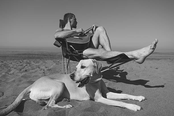 A man sitting in the camp chair on the beach.He is playing a guitar while his dog sits next to him