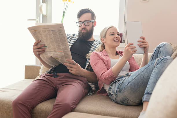 Reading news Young couple in love sitting on a couch in their apartment next to the window, enjoying their free time, reading newspaper and surfing the web on a tablet computer. Lens flare effect on window reading newspaper stock pictures, royalty-free photos & images