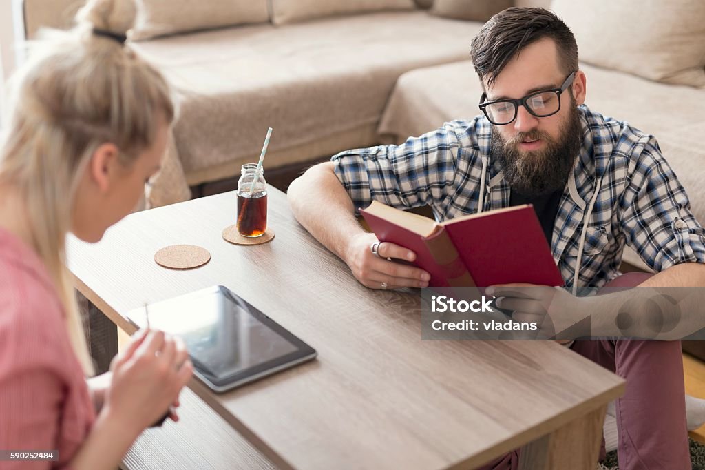 Reading relaxation Young couple in love sitting on the floor next to a table in a living room of their apartment, reading and relaxing on their leisure time. Lens flare effect on the window Adult Stock Photo