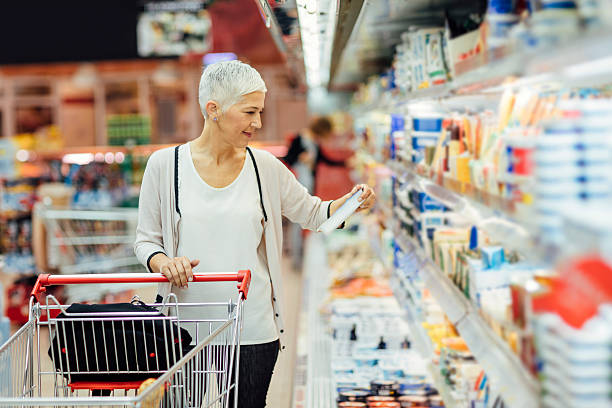 Mature Woman Groceries Shopping. Mature smiling woman shopping in local supermarket. She is shopping groceries. Standing by produce stand and reading nutrition label on dairy product. Choosing cheese. farmers market healthy lifestyle choice people stock pictures, royalty-free photos & images