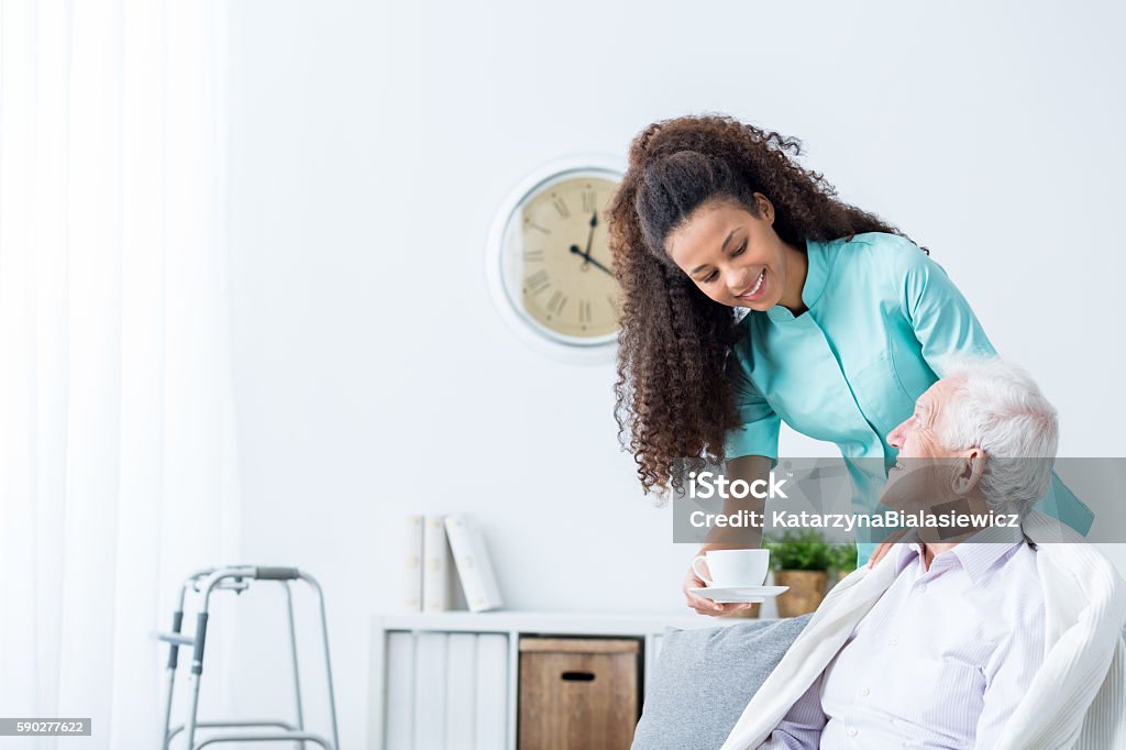 Female caregiver serving afternoon tea to patient Pretty young caregiver serving afternoon cup of tea to older happy man Home Caregiver Stock Photo