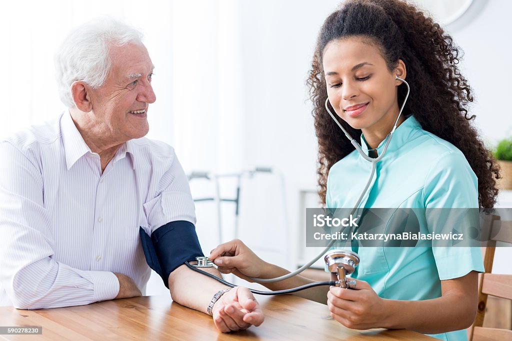 Professional nurse checking patient's blood pressure Young happy professional nurse checking senior man's blood pressure Assistant Stock Photo