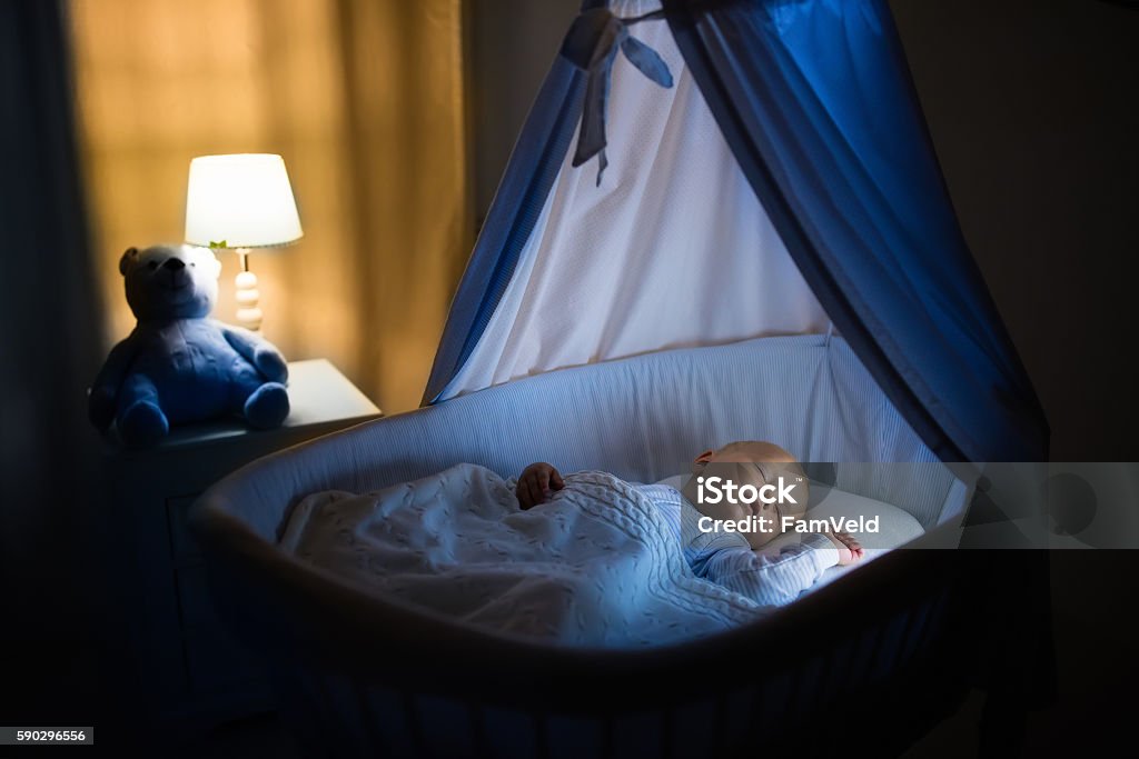 Cute baby boy sleeping at night Adorable baby sleeping in blue bassinet with canopy at night. Little boy in pajamas taking a nap in dark room with crib, lamp and toy bear. Bed time for kids. Bedroom and nursery interior. Sleeping Stock Photo