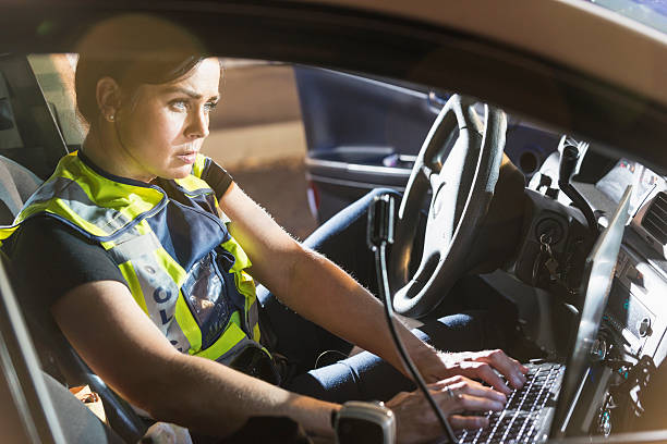Policewoman sitting in patrol car using computer A female police officer sitting in her police car, using the computer. It is nighttime and the emergency lights on her vehicle are flashing. female police officer stock pictures, royalty-free photos & images