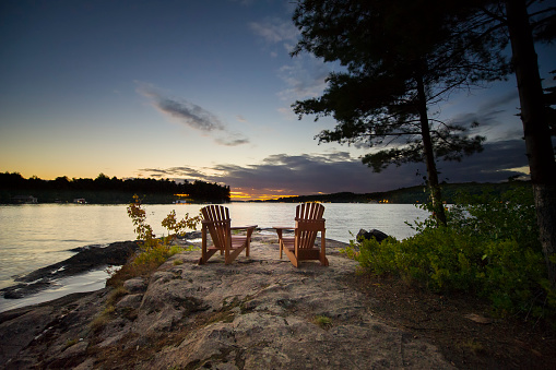 Two Muskoka chairs sitting on a rock facing a calm lake. Across the calm water is a white cottage nestled among green trees. Time of the day is sunset.