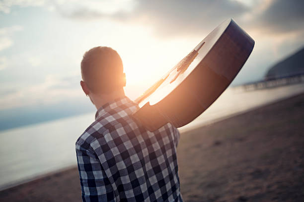 Rear View Of Man with Acoustic Guitar At Beach how relaxing and looking at the sunrise.
