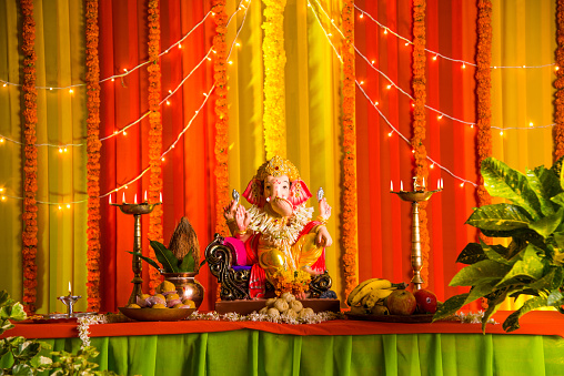 A clay statue of an Indian god Lord Ganesha on ganesh festival over table decorated with garland and lighting series, fruits and indian traditional sweets are offered