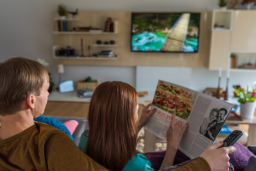 Man sitting on sofa and watching television show while woman reading magazine.