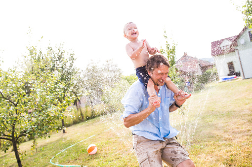 Father carrying his son on his shoulders at the sprinkler, fun in garden, sunny summer day, back yard