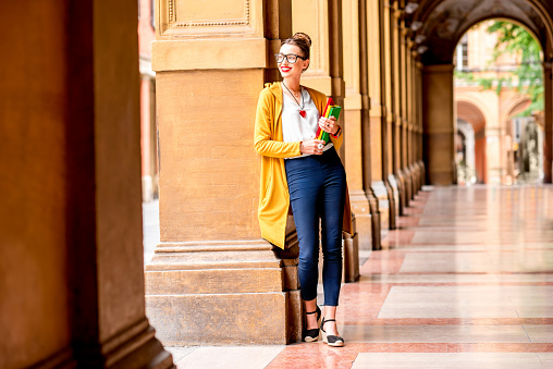Young female student dressed casually standing with books in the famous arched galleries in Bologna city in Italy. Bologna is student city and home to the oldest university in the world
