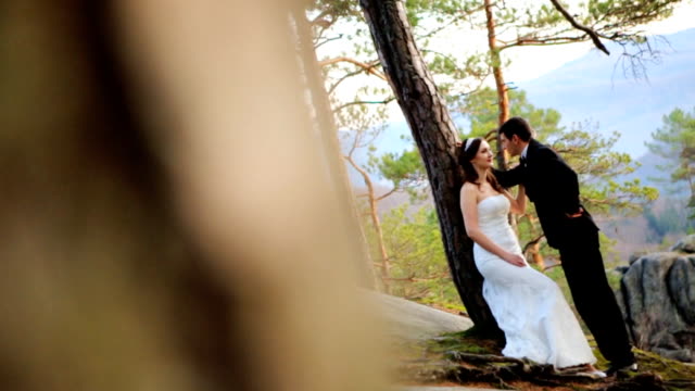 Happy brunette bride leaning against a tree looking at her handsome groom in pine forest