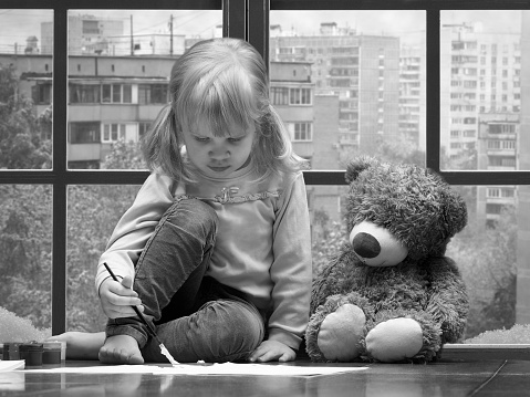 Little girl draws paints and brush. A huge window behind the glass city, snow, winter. toy teddy bear sitting next to. Black and white image