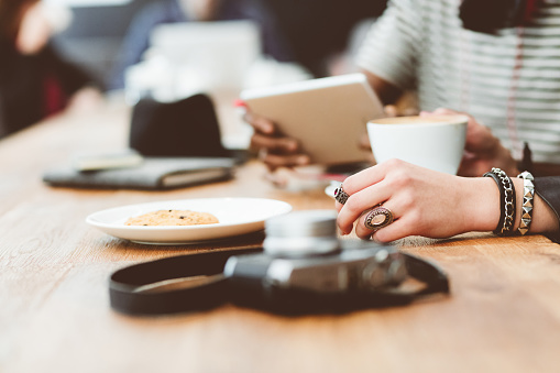 Young people sitting at the table in a coffee shop. Afro american guy holding a digital tablet, woman drinking coffee. Close up of hands, unrecognizable people.