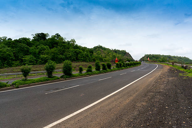 Long stretch of empty road bordered with tree filled hills and under beautiful monsoon skies. Showing a great way to explore india via the well developed road network