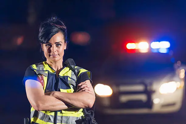 A policewoman wearing a safety vest standing in the street at night. In the background, her police car is out of focus, with lights illuminated. She is smiling at the camera with her arms crossed.