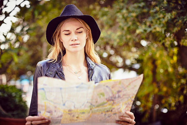 istock Beautiful young woman wearing cap while reading map against tree 591811010