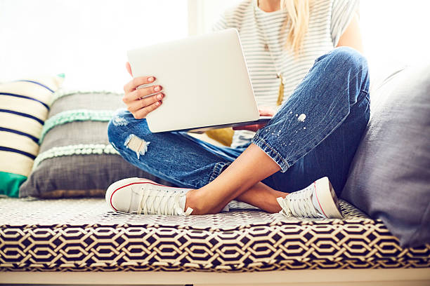 Low section of trendy woman using laptop. Female hipster is sitting with legs crossed on window seat. Woman is wearing blue jeans and canvas shoes at home.