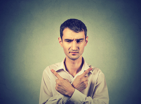 Confused young man pointing in two different directions, hesitant to make decision isolated on gray background. Human emotion, facial expression reaction