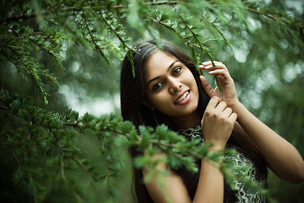 Beautiful girl between nature full of branches of pine tree. Outdoor image of beautiful, happy late teen urban girl in nature at day time looking at camera with toothy smile behind the green branches of pine tree. One person, horizontal composition with selective focus and copy space. teenage girls pretty smile looking at camera waist up stock pictures, royalty-free photos & images