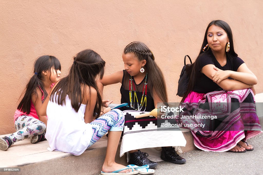 Santa Fe 2016 Indian Market: Native American Family Santa Fe, NM, USA - August 20, 2016: A Native American family sits near an adobe wall at the 2016 Santa Fe Indian Market. One of the girls wears a traditional native dress. The market, now in its 95th year, is spread out all around the historic Santa Fe Plaza, showcasing North American Indigenous arts and culture. About 1,000 artists from 220 tribes participate in the two-day event. Child Stock Photo