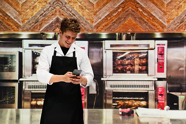 sorridente chef em cozinha usando telefone - chicken rotisserie barbecue grill roast chicken - fotografias e filmes do acervo
