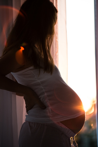 Pregnant mid adult Caucasian woman supporting her back with hand, standing by the window.