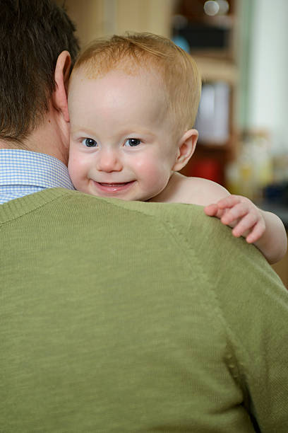 A baby girl holds on to daddy's shoulder and smiles. Photo with editorial space