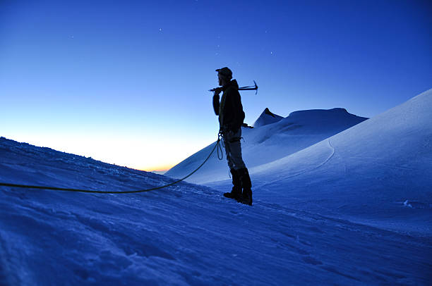 Hiker with an ice axe resting and watching the dawn at an elevation of 3800m above sea level in the Swiss Alps near the Summit of Alphubel (4206m). Allalinhorn in the background. Focus on background.