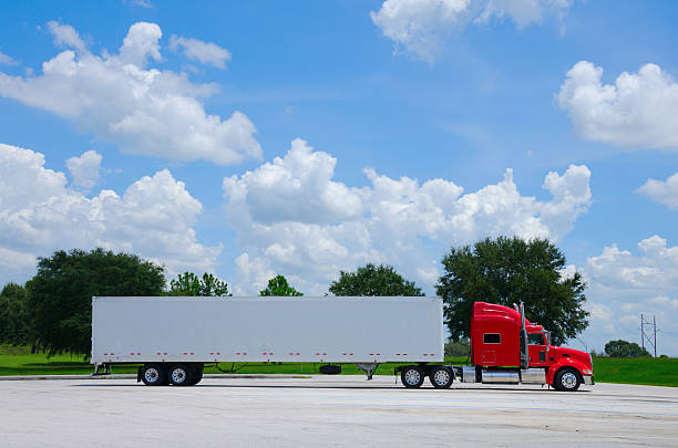 Clean shiny red 16-wheel semi tractor truck with a clean white cargo container trailer against a simple colorful background of trees and blue sky