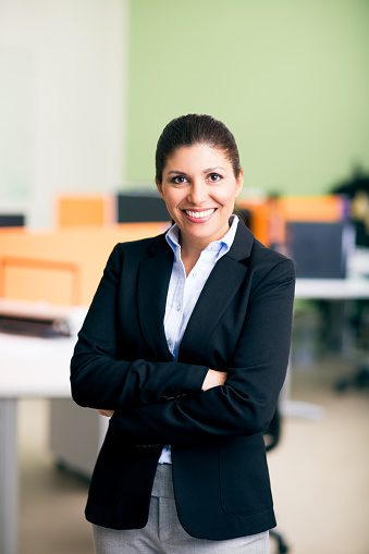 A latin woman office administrator standing with arms crossed and smiling at the camera in a vertical medium shot indoors.