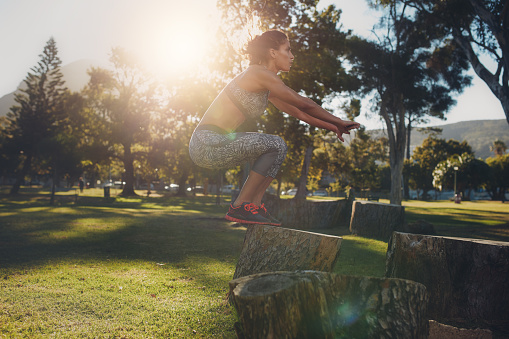 Shot of a young woman jumping onto wooden log at the park. Sportswoman doing exercise in nature on a sunny day.