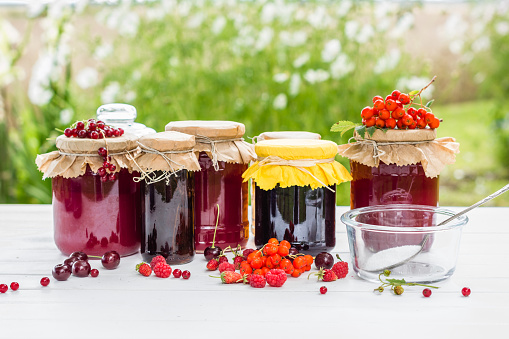 Fruit preserves and raw strawberries , cherries , rowanberries and red currants berries on a white wooden table, on the nature background, organic meal and dessert concept