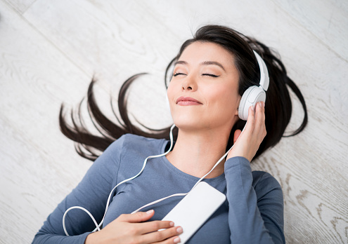 Happy Latin American woman listening to music at home on her cell phone lying on the floor with headphones and eyes closed
