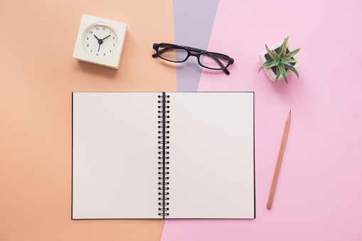 Top view of open empty spiral notebook with pen, eyeglasses, clock, and plant on pastel colorful background