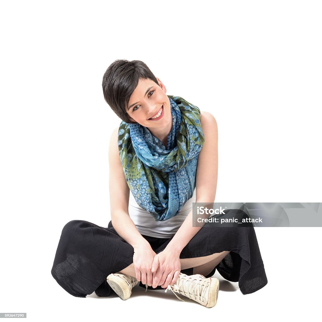 Charming girl sitting cross-legged smiling at camera with slanted head Charming pretty girl sitting cross-legged smiling at camera with slanted head over white studio background Young Women Stock Photo