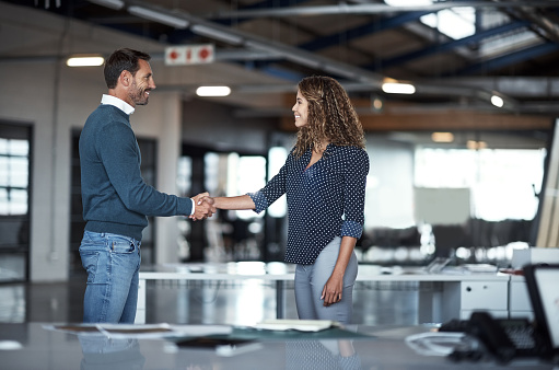 Shot of two coworkers shaking hands together while standing in a large modern office