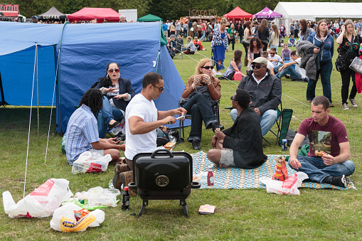 Liverpool, United Kingdom - June 22, 2013: Group of people, mixed age and race, at an outdoor music annual local music and craft festival, Larks in the Parks, held in Liverpool, sitting a circle around ground blue and white cloth, cooking and eating BBQ, with a scattered crowd and tents in the background. 