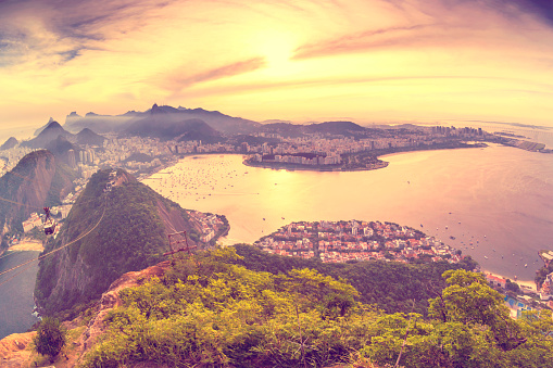 Aerial view of Rio de Janeiro at sunset.  The beach is visible in the bottom-right section of the photo.There is a small forested area in the middle of the photo.  Buildings surround the beach and forest area, extending toward the mountains in the background.  The mountains are blanketed by mist that partially obscures them, but the Christ the Redeemer statue is visible above the mist.  The sunlight adds a warm glow to the photo, and the area behind the mountains is golden.