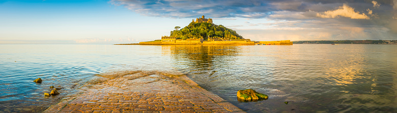 Gentle ocean tide ebbing across the iconic cobbled causeway as warm summer sunlight illuminates the historic chapel, hamlet and harbour of St. Michael's Mount, Marazion, Cornwall, UK.