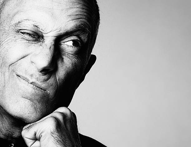 Senior man grimaces cynically looking to the side A good-looking older man resting his chin on his hand grimaces, screwing up one eye and looking to the side, where there is copy space, in irritated disbelief. Black and white image. skeptical look stock pictures, royalty-free photos & images