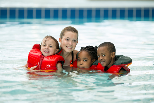 A multi-ethnic group of elementary age children are taking a swim class together at the public pool. They are smiling and looking at the camera.