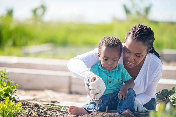 A mother is teaching her toddler son how to plant food in the vegetable garden. They are spending mother's day out in the sun gardening together.