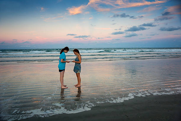 Teen girls with smartphones on ocean beach at sunset Two teenaged barefoot Caucasian girls face each other while they text or send a photo on their smartphones  on the beach at dusk on vacation in Cocoa Beach, Florida, USA cocoa beach stock pictures, royalty-free photos & images