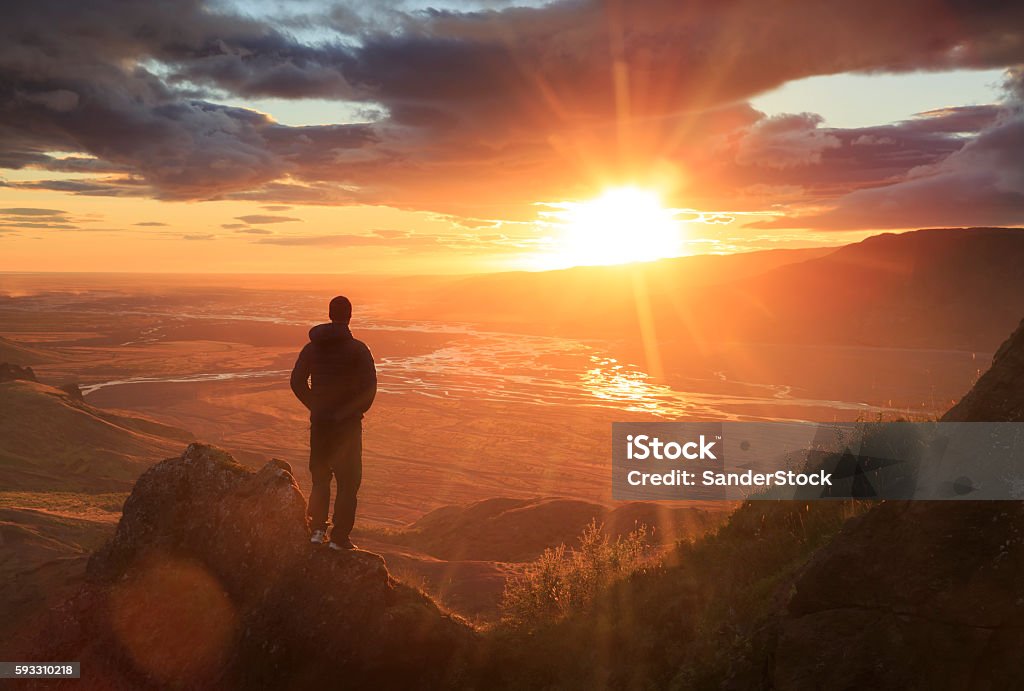 Looking at the sun Man standing on a ledge of a mountain, enjoying the sunset over a river valley in Thorsmork, Iceland. With lens flare. Mountain Stock Photo
