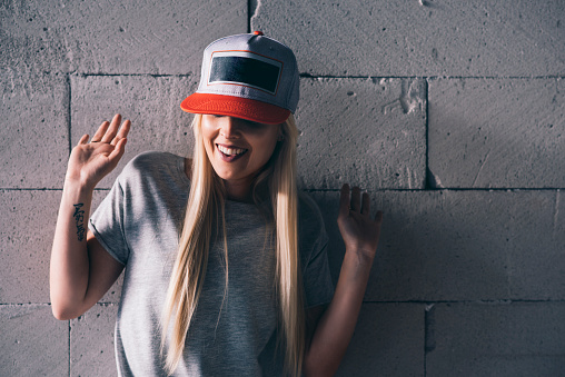 Young woman with cap on her head is standing in front of white wall. She is looking down.