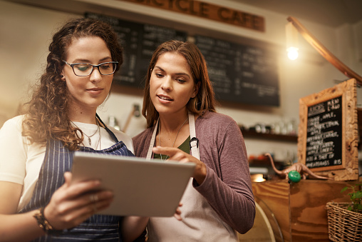 Cropped shot of two young women looking at a tablet while sitting in their bakery