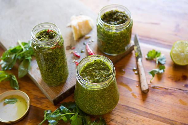 A horizontal composition of three glass jars filled with fresh homemade basil and coriander pesto, displayed on a wooden background with other ingredients including chilli peppers, parmesan cheese and lime. 