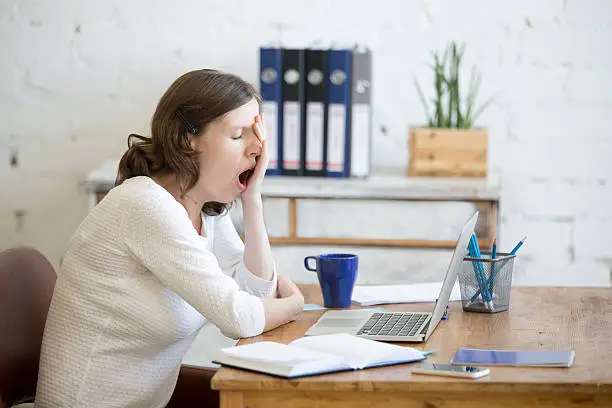 Portrait of young woman sitting at table in front of laptop, sleepy, tired, overworked, lazy to work. Attractive business woman yawning in home office relaxing or bored after work on laptop computer