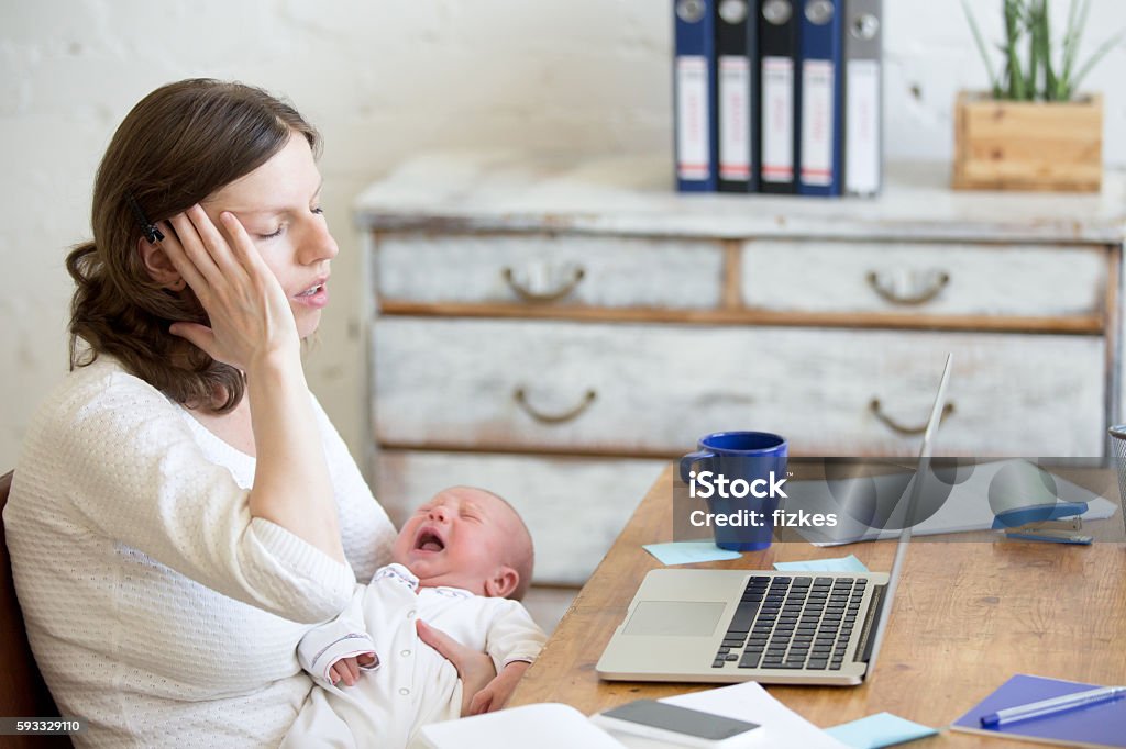 Portrait of young business mom having headache because of crying Young businesswoman holding newborn crying babe while sitting with laptop and touching head with pained expression. Career mom suffering from migraine after working on pc and nursing crying child Mother Stock Photo