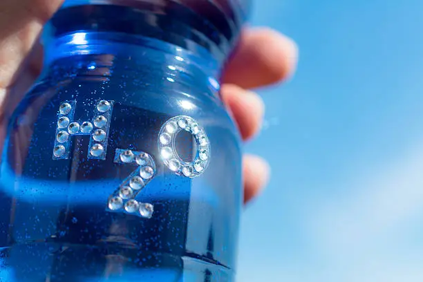 Close up hand holding Bottle of water with rhinestone sparkle  text H20 the chemical name of water against a blue sky